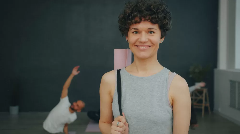 A woman smiling with a rolled mat over her shoulder while another practices a side stretch behind her