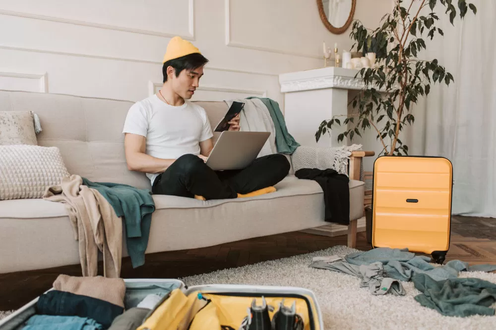 A traveller sitting cross-legged on a sofa with a laptop and phone, a suitcase open on the floor beside him