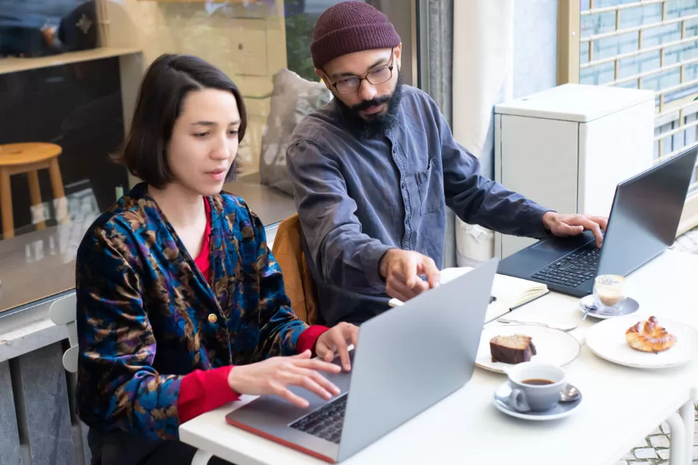 Two people working at laptops in a café window seat, coffee, cake and pastries on the table between them