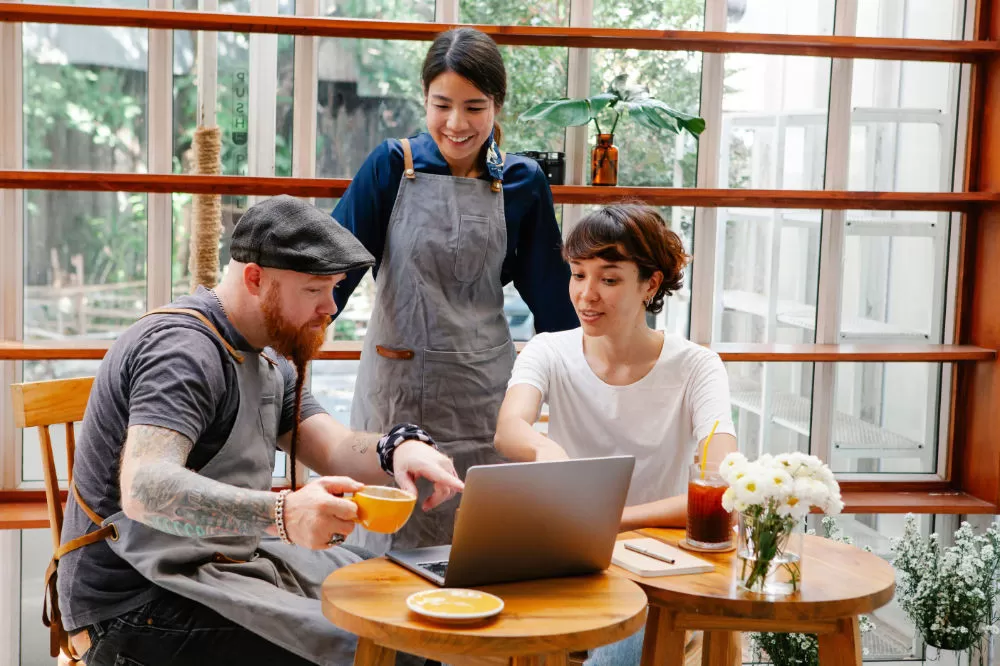 A barista in an apron leaning over two seated colleagues looking at a laptop in a plant-filled café