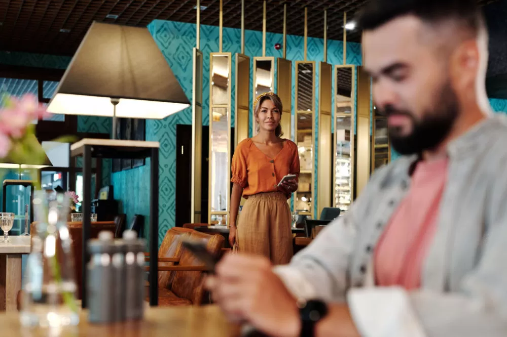 A host crossing a hotel bar toward a guest who is checking his phone at the counter in the foreground