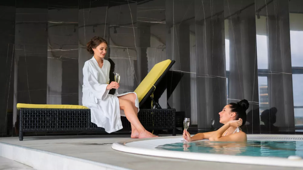 Two women with champagne at a spa, one in a robe on a lounger and one resting in the hot tub