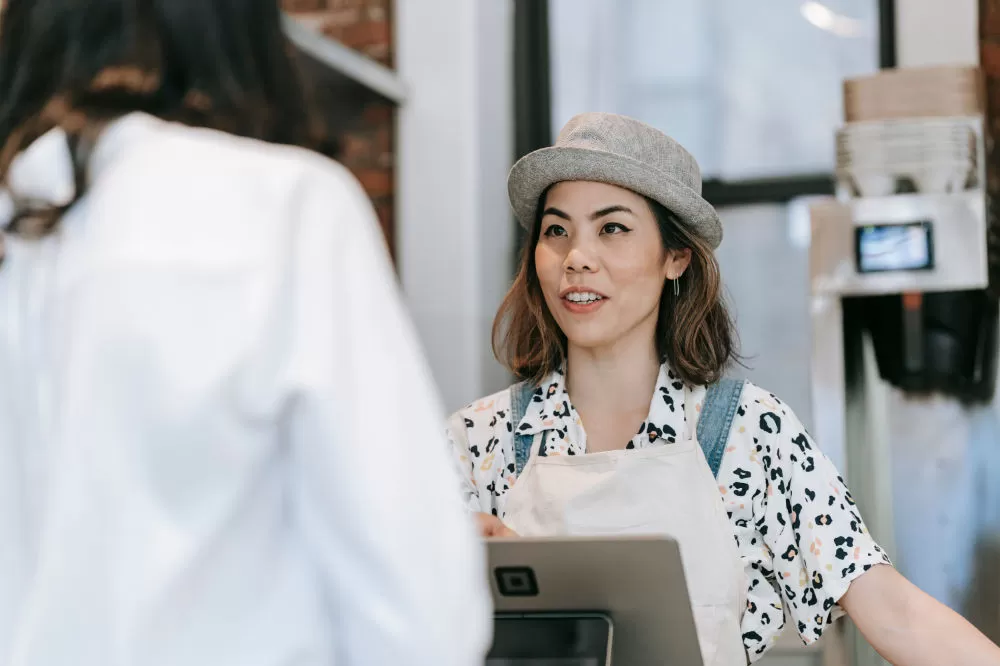 A café worker in an apron and flat cap taking a customer’s order at the counter beside the till