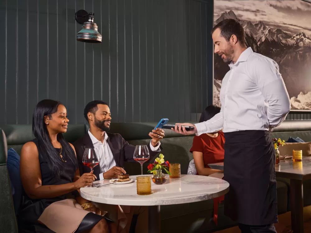 A server holding out a card terminal as a guest pays by phone at a restaurant banquette, wine on the table