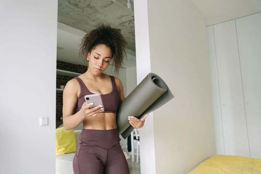 A woman in workout clothes checking her phone at home, a rolled yoga mat tucked under her arm