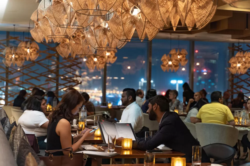 A full restaurant dining room at night, woven pendant lights overhead and a lit city skyline through the windows