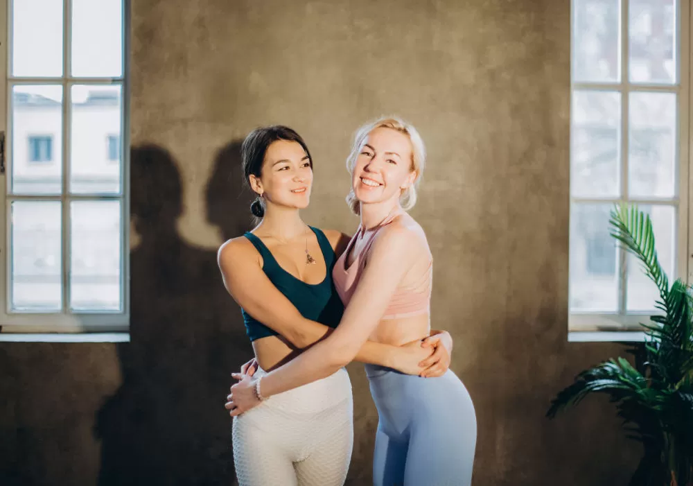 Two women in activewear standing arm in arm, smiling, in a daylit studio between two tall windows