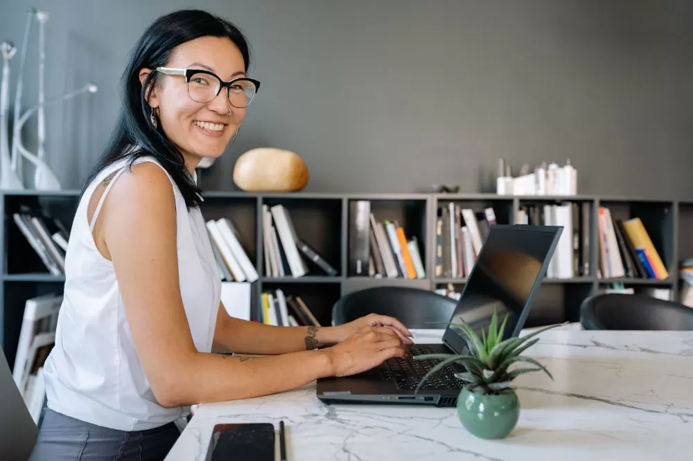 A woman at a marble desk turning from her laptop to smile, bookshelves filling the wall behind her